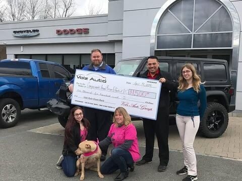 People and a dog posing outside a dealership with a donation check