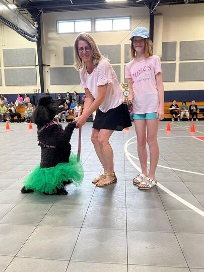 Woman and child posing with a dressed-up poodle holding a trophy.