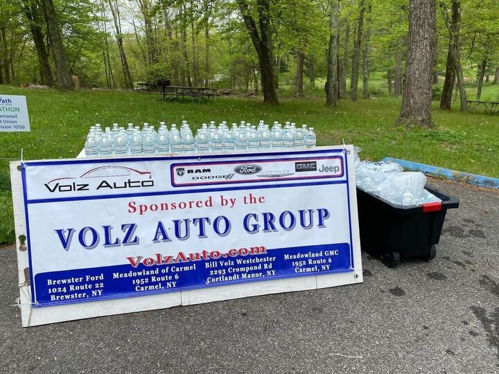 A 'Volz Auto Group' sponsor banner with Ram, Dodge, Ford, Chrysler, GMC, and Jeep logos. Many water bottles are on and near tables in a park.