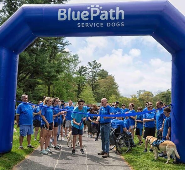 People in blue shirts and a service dog stand at a blue 'BluePath Service Dogs' inflatable arch at a run.#