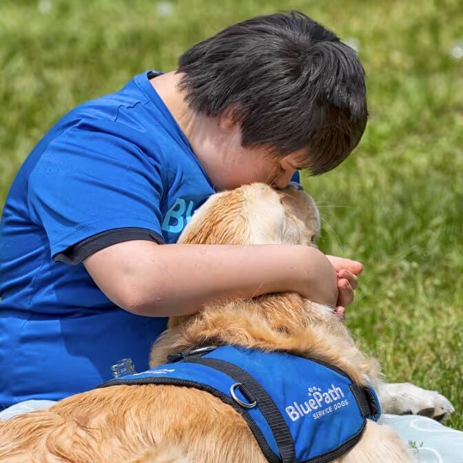 A person in a blue shirt with dark hair embraces a Golden Retriever wearing a blue 'BluePath Service Dogs' harness.